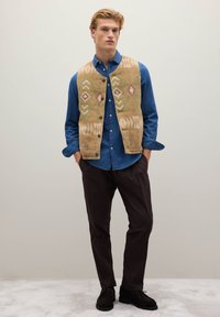 Beige gemusterte Weste mit geometrischen Mustern über einem blauen Hemd mit Knöpfen, kombiniert mit dunkelbraunen Hosen und schwarzen Schuhen.