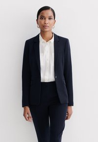 Completo da uomo blu navy con blazer su misura dotato di due tasche, abbinato a una blusa bianca e pantaloni su misura coordinati.