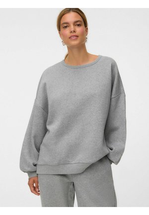 Femme portant un sweat-shirt ample gris clair et un pantalon de jogging assorti, debout devant un fond clair uni.