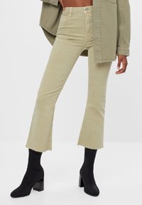 Pantalones de pana acampanados en verde claro con dobladillo raw, combinados con botas negras de tacón cuadrado. La tela tiene un acabado texturizado.