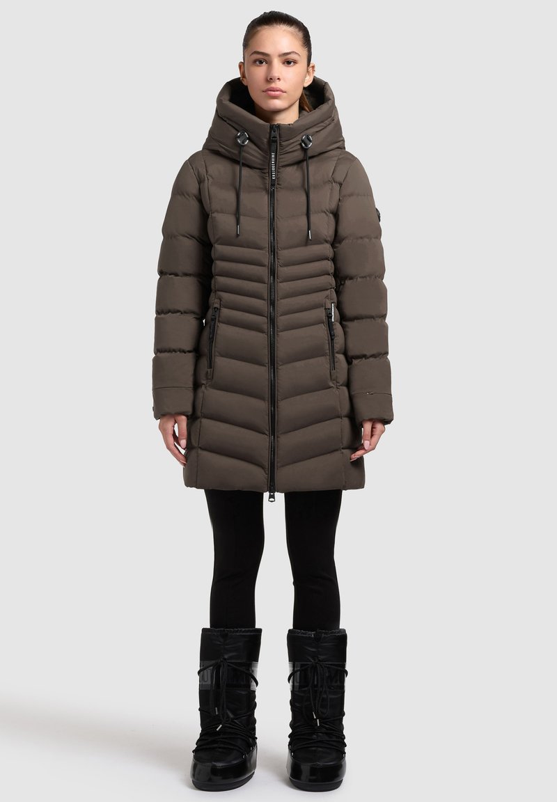 Puffer coat in dark brown, featuring a zip front, ribbed panels, side pockets, and a drawstring hood. Worn with black leggings and boots.