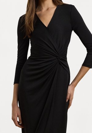 Musta mekko, valmistettu pehmeästä, joustavasta kankaasta, jossa syvä V-pääntie, pitkät hihat ja vyötäröllä kiertynyt solmup detaili.