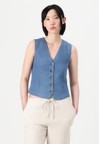 Blå denimærmeløs vest med V-hals og fem knapper foran. Bæres over lys beige bukser med afslappet pasform.