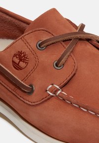 Chaussure bateau en cuir marron avec détails à lacets, coutures blanches et logo d'arbre embossé sur le côté près des œillets.