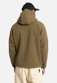 Felpa con cappuccio marrone in pile con superficie strutturata e un grande cappuccio. Un dettaglio ricamato a forma di cactus è visibile su pantaloni color crema.