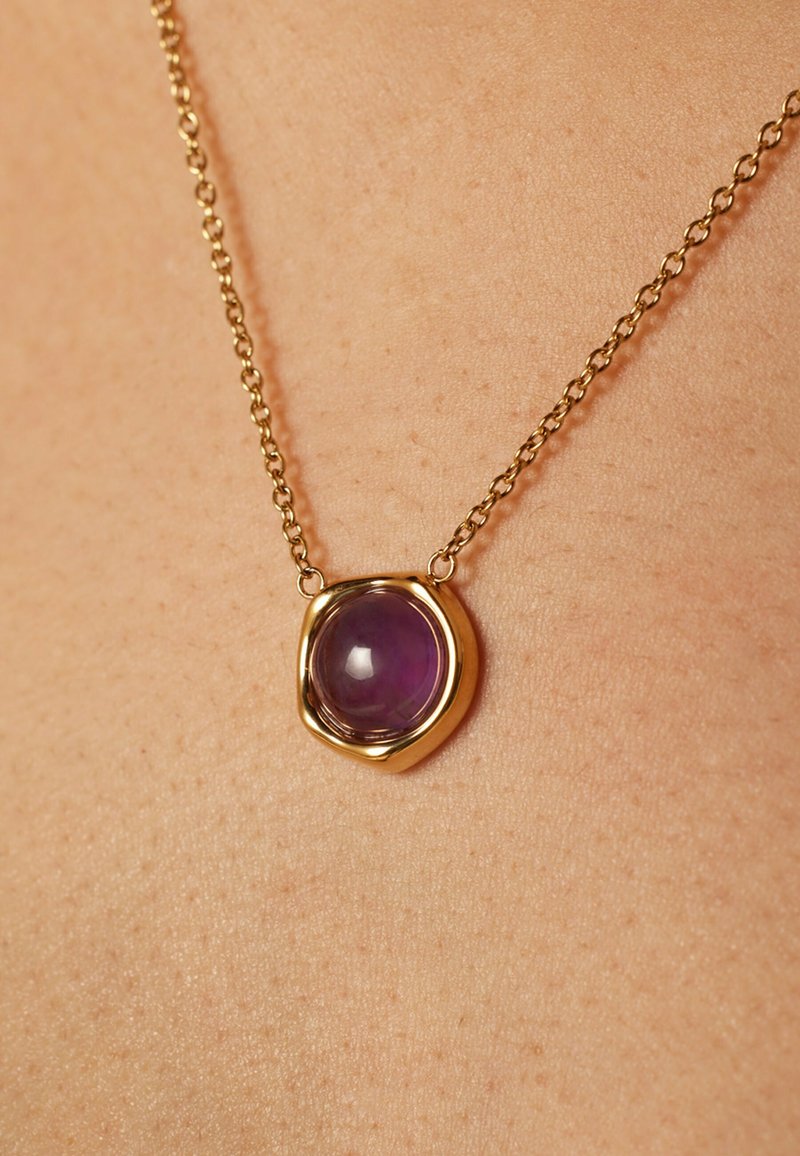 Gouden halsketting met een afgeronde paarse edelsteen hanger in een organic-vormige zetting, hangend aan een delicate ketting.