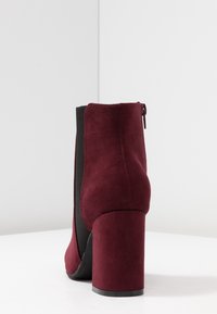 Botín burdeos de ante con acabado liso, tacón alto ancho y paneles elásticos laterales. Cuenta con cierre de cremallera en la parte trasera.