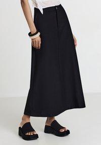 Schwarzer Maxikleid aus glattem Stoff, mit einem Frontknopfverschluss, seitlichen Taschen und einer geraden Silhouette, kombiniert mit klobigen schwarzen Sandalen.