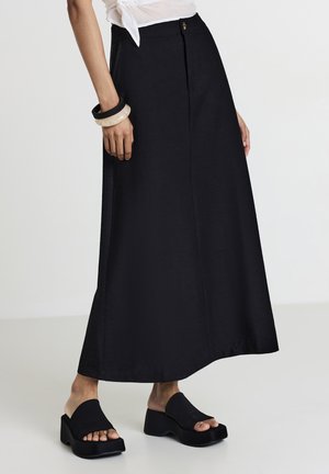 Gonna maxi nera realizzata in tessuto morbido, con chiusura anteriore a bottoni, tasche laterali e silhouette dritta, abbinata a sandali neri spessi.