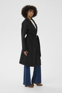 Černý kabát s páskem, strukturovaným límcem a dlouhými rukávy, vyrobený z hladkého materiálu. Nesený přes bílý top a kombinovaný s rozšířenými džíny.