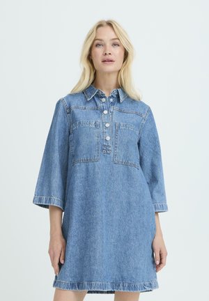 Femme blonde portant une robe en jean bleue avec des manches trois-quarts, des poches avant et des boutons argentés sur la patte de col, debout devant un fond blanc.