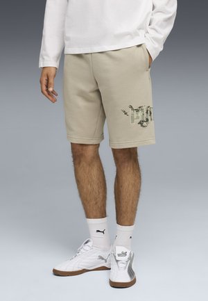 Man wearing beige Puma shorts, white Puma socks, and white Puma sneakers, hand in pocket, against neutral gradient background.