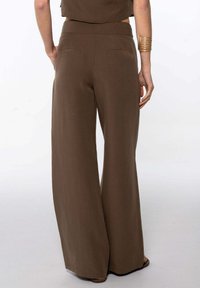 Femme portant un pantalon large taille haute marron et un haut assorti, avec un bracelet en or au poignet droit, se tenant sur un fond clair.