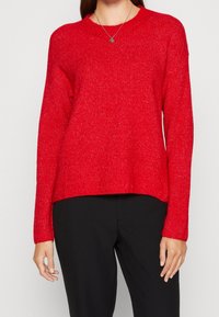 Pull en tricot rouge avec un col rond, des manches longues et un ourlet légèrement court. Le tissu a une finition texturée et scintillante.