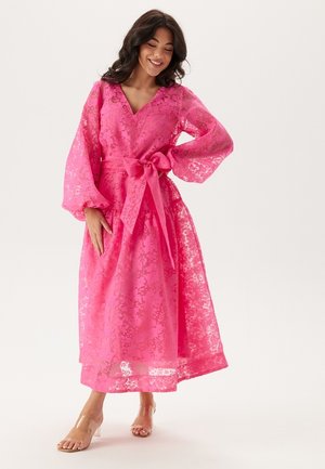 Long pink lace dress featuring a v-neck, flowy balloon sleeves, a tied waist, and a full skirt with a sheer patterned overlay.