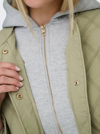 Nærbillede af en person iført en lysegrå lynlåshættetrøje under en quiltet olivengrøn jakke med guldknapper.