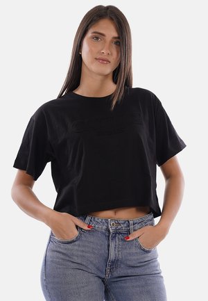 Giovane donna con lunghi capelli scuri indossa una maglietta nera corta e jeans blu a vita alta, posando con le mani nelle tasche su sfondo bianco.