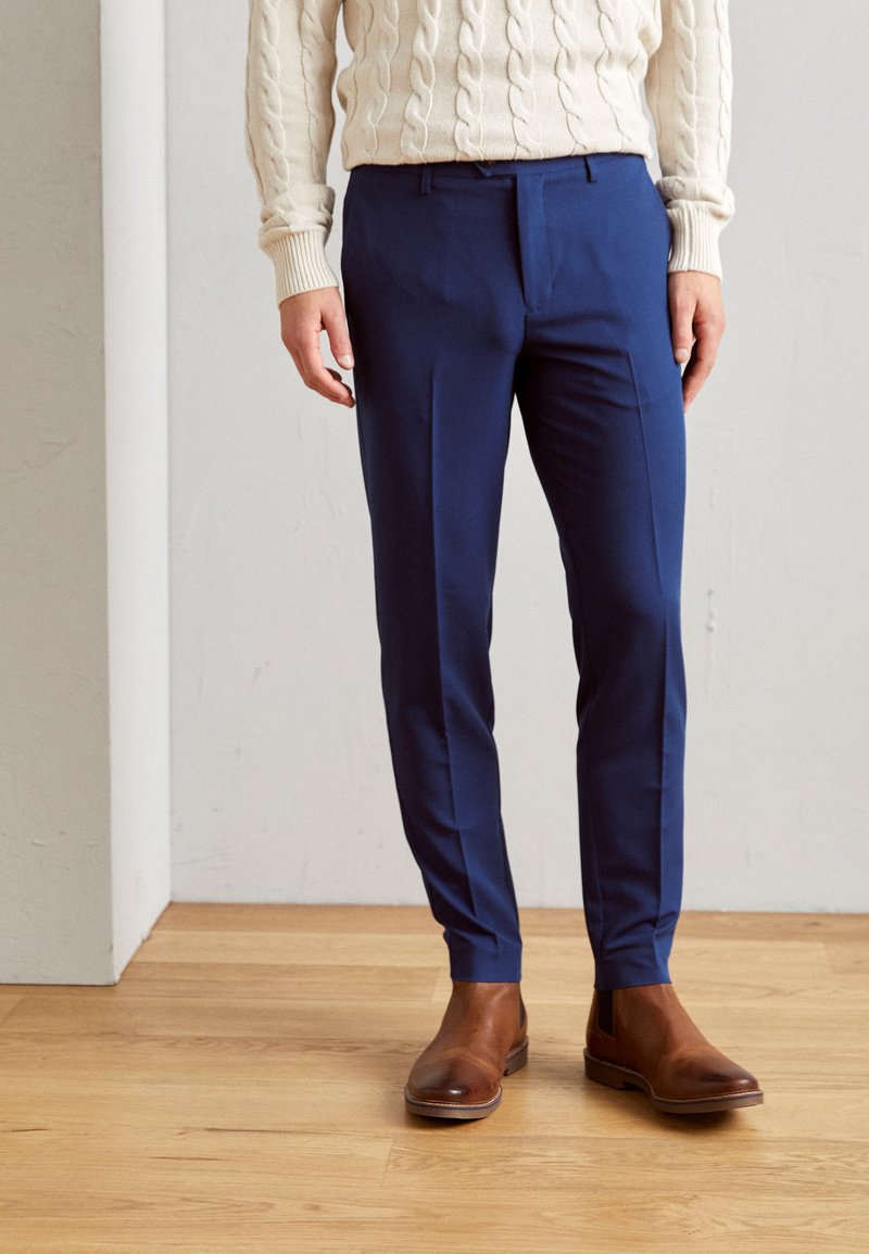Navy blue tailored trousers with a slim fit, featuring smooth fabric and subtle front pleats, paired with brown leather chelsea boots.