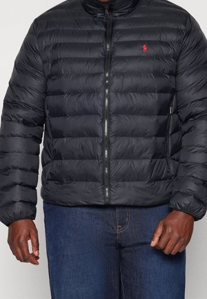 Mann trägt eine schwarze gesteppte Jacke mit Reißverschluss und rotem Logo auf der Brust sowie blaue Jeans, steht mit entspannten Armen an den Seiten.