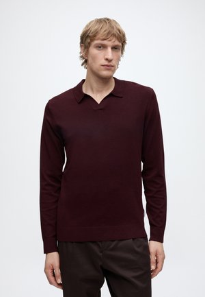 Homme blond portant un pull bordeaux foncé à manches longues avec un col et un pantalon foncé, debout devant un fond clair uni.