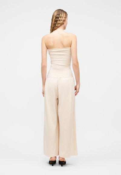 Γυναίκα με μακριά κυματιστά μαλλιά, φορώντας ένα μπεζ τοπ χωρίς τιράντες και μπεζ παντελόνι με φαρδιά πλάγια, στέκεται με την πλάτη προς τα εμπρός φορώντας μαύρα τακούνια.