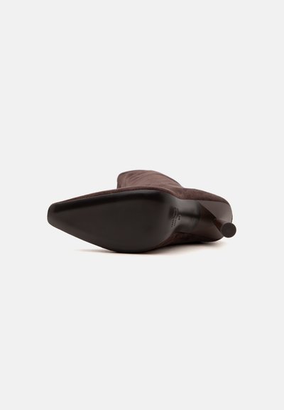 Botte stiletto en daim marron à bout pointu posée sur le côté, montrant la semelle et le talon sur un fond uni.