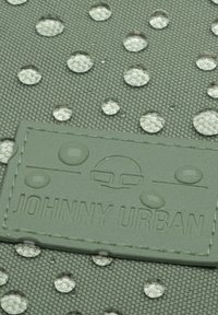 Gros plan d'un tissu vert avec des gouttes d'eau et une étiquette cousue indiquant "Johnny Urban" avec un logo visage minimaliste.