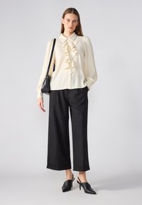 Blusa blanca texturizada con un frente con volantes y cierre de botones, combinada con pantalones de pierna ancha negros de talle alto y tacones negros tipo slingback.