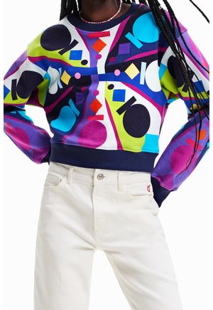 Svítivě vzorovaná mikina s výraznými tvary v modré, zelené, fialové a černé barvě; kombinovaná s bílými kalhotami a námořnicky modrými žebrovanými manžetami.