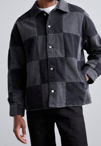 Schwarz-grau kariertes Jeansjacke mit Kragen, silbernen Druckknöpfen und ausgefransten Kanten. Kombiniert mit schwarzen Hosen.