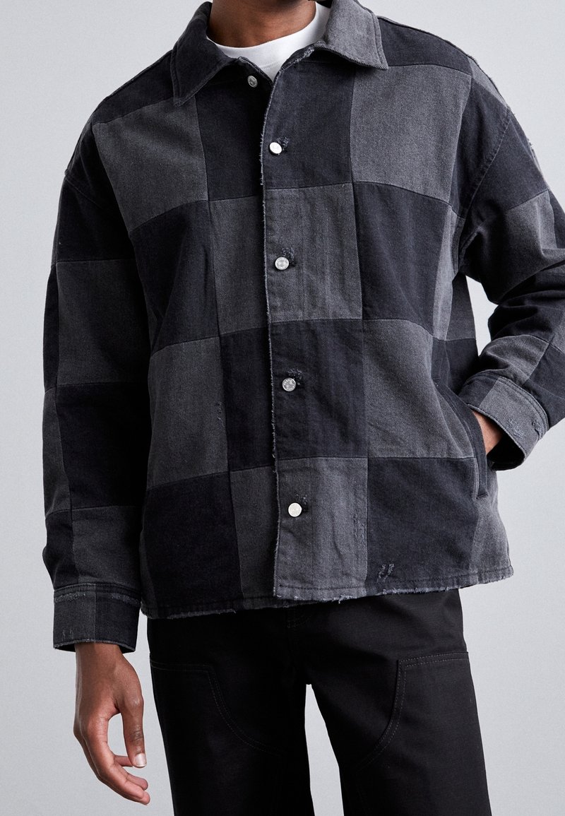 Schwarz-grau kariertes Jeansjacke mit Kragen, silbernen Druckknöpfen und ausgefransten Kanten. Kombiniert mit schwarzen Hosen.