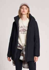 Młoda kobieta nosząca beżową czapkę, kremową bluzę z grafikiem, czarną kurtkę zapinaną na zamek, czarne legginsy oraz koszulę w kratę przewiązaną wokół talii.