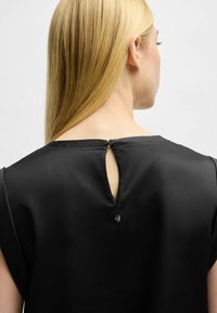 Blusa nera con scollatura rotonda e una piccola apertura a forma di chiave sul retro, con un dettaglio di bottone decorativo. Tessuto liscio e testurizzato.