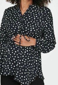Blusa nera con pois bianchi, maniche lunghe, scollatura con bottoni e dettaglio con laccio in vita, progettata per l'abbigliamento maternità.