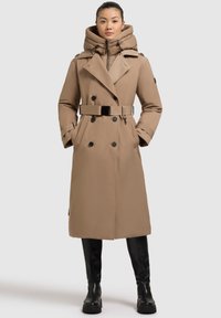 Trench coat beige con abbottonatura doppia, cappuccio grande e cintura. Realizzato in un tessuto liscio, dotato di tasche laterali e bottoni neri.