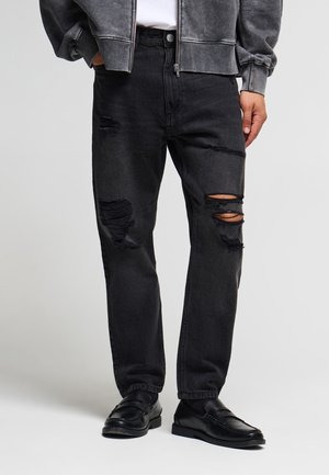Persona che indossa jeans neri strappati, mocassini neri e una giacca grigia con cerniera, con le mani nelle tasche su uno sfondo uniforme.