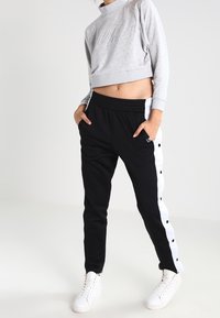 Grijze cropped sweatshirt met reliëflogo, gecombineerd met zwarte joggers met witte zijknopen. Beide items hebben een relaxed fit.