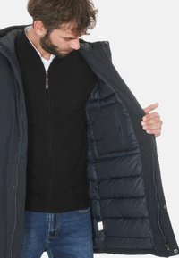 Giacca trapuntata blu navy con cappuccio, aperta per mostrare l'imbottitura e una tasca laterale. Indossata sopra un maglione nero con zip e jeans blu.