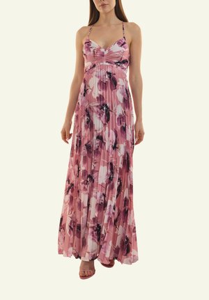 Frau trägt ein rosa, plissiertes Maxikleid mit lila und weißen Blumenmustern und dünnen Schulterriemen, kombiniert mit passenden offenen High Heels.