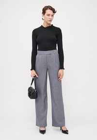 Zwart lange mouwtop met een gerimpelde halslijn; grijze wide-leg broek; houdt een kleine zwarte handtas vast; zwarte hakken met een spitse neus.