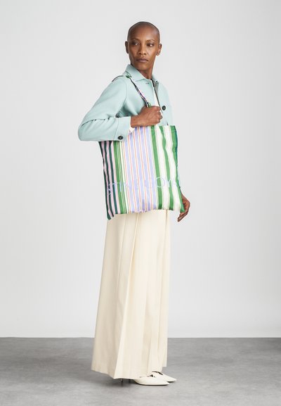 Tote-väska med vertikala ränder i grön, rosa och vit. Har ett mjukt tyg och axelremmar. Modellen bär en ljusblå jacka och krämfärgade byxor.