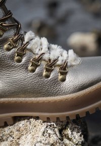 Botín de cuero dorado metálico con un forro blanco esponjoso, cordones marrones y herrajes metálicos dorados. Presenta una suela de goma gruesa y texturizada.