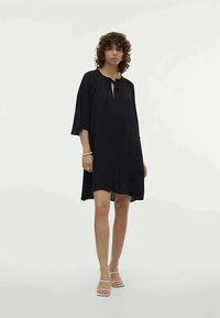 Juodas trumpas suknelė su V formos iškirpte ir auriu detaliu, su trimis ketvirtadaliais rankovėmis ir laisvu pavyzdžiu. Lygus paviršius, iki kelių ilgio.