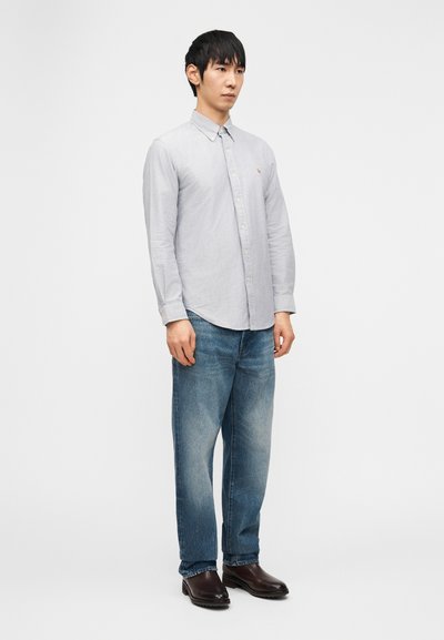 Άνδρας που στέκεται, φοράει ανοιχτό γκρι πουκάμισο με κουμπιά, ξεθωριασμένο μπλε τζιν και σκούρα καφέ δερμάτινα μποτάκια, μπροστά σε απλό λευκό φόντο.