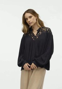 Schwarze Bluse mit einer Spitzenblende und gefältelter Vorderseite. Sie hat lange, luftige Ärmel und Rüschensaum. Getragen mit leichten beigen Hosen.