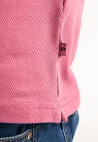 Polo rose en tissu texturé, avec une petite étiquette logo sur la couture latérale et des bords finis. Porté par-dessus un denim foncé.