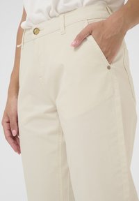 Jasno-beżowe bawełniane spodnie o slim fit, z bocznymi kieszeniami i mosiężnym guzikiem w pasie. Gładka tekstura i prosty design.