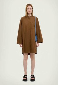 Braunes, übergroßes Hemdkleid mit langen Ärmeln, einem Mandarin-Kragen und einer Brusttasche. Kombiniert mit schwarzen Plateausandalen und einer blauen Handtasche.