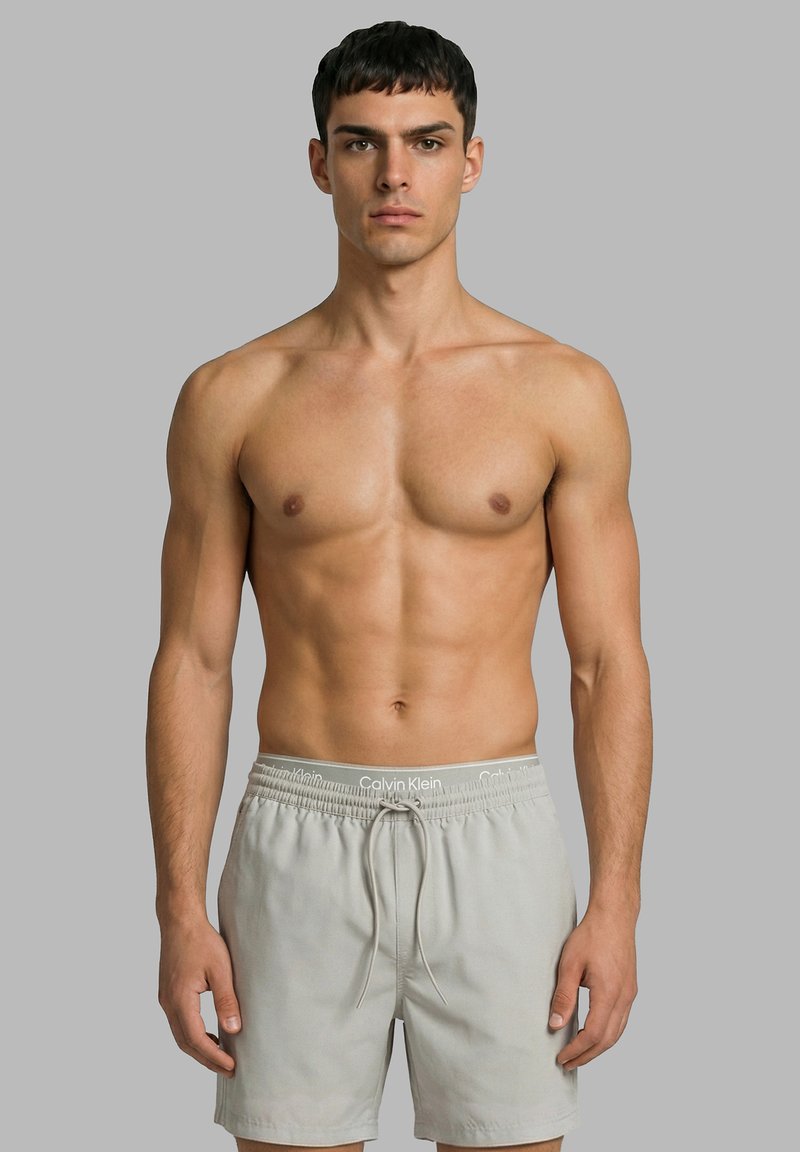 Jonge man met kort donker haar, gekleed in lichtgrijze zwembroek met zichtbare Calvin Klein tailleband, staand tegen een grijze achtergrond.
