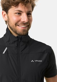 Gilet nero con chiusura a zip, collo alto e accenti riflettenti. Presenta il logo VAUDE in posizione prominente sul lato sinistro. Maniche corte visibili.